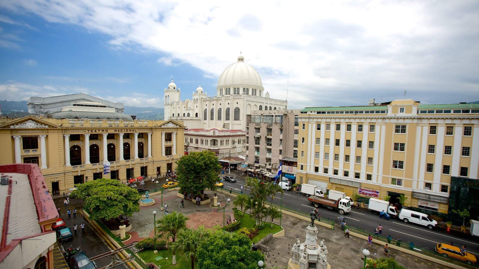 Aerial view of a city plaza with Teatro Nacional, cathedral dome, street traffic, and pedestrians in El Salvador.