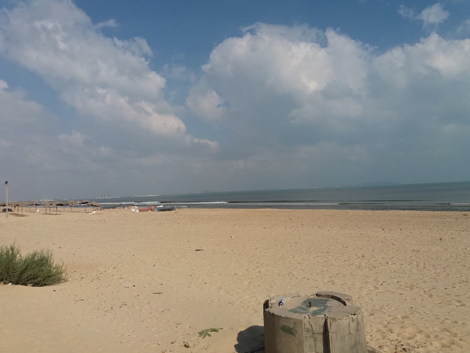A wide sandy beach scene with calm ocean waters, distant umbrellas, and a blue sky with clouds, featuring a concrete structure in the foreground.