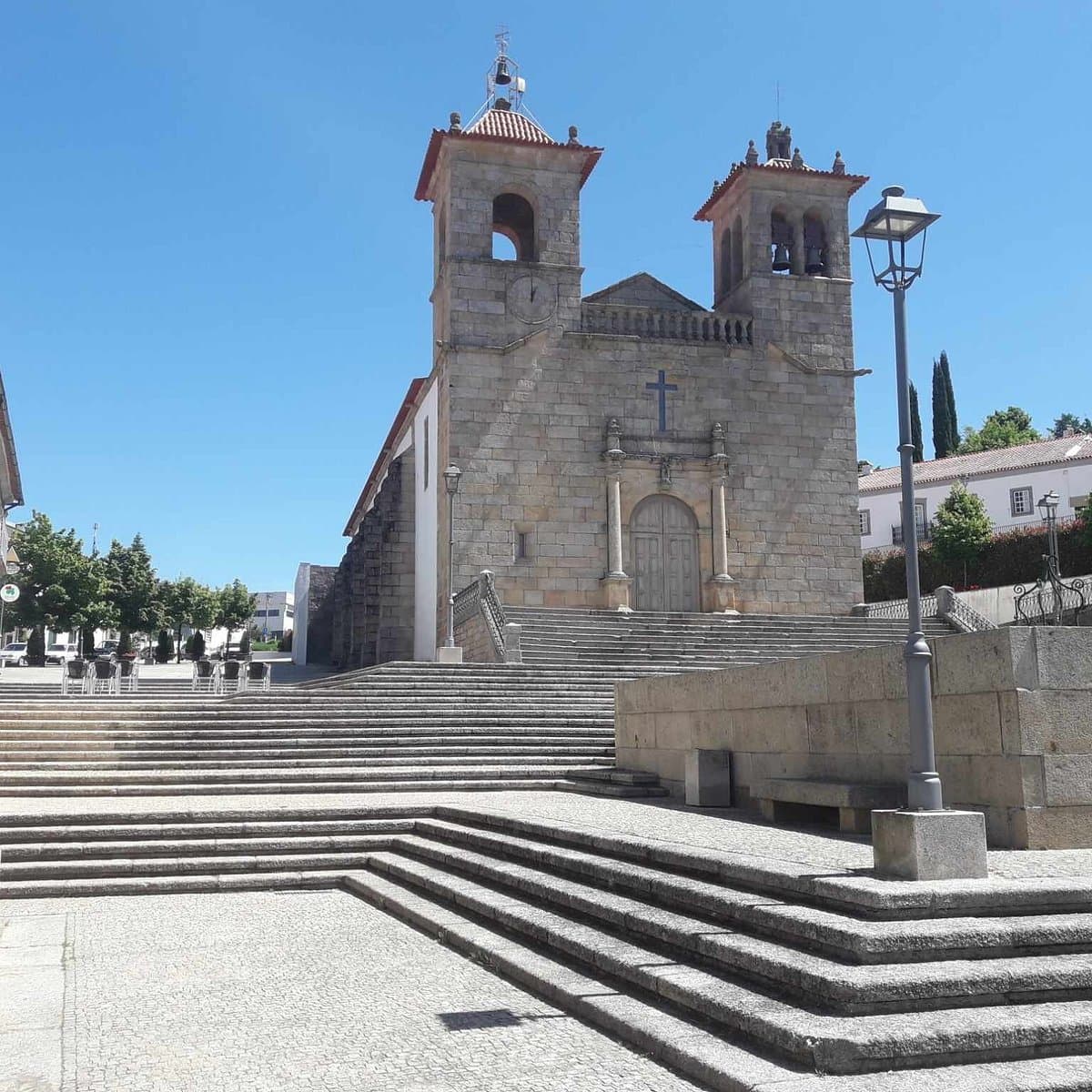 Sunlit stone church facade with twin bell towers and wide steps in a plaza, likely in Vimioso, Portugal.