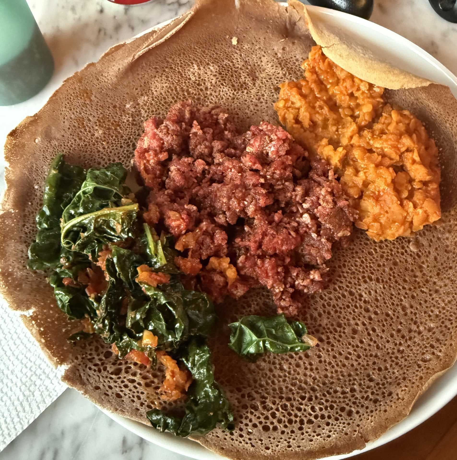 Overhead photograph of an Ethiopian platter with injera, red meat stew, orange lentil stew, and green leafy vegetables.