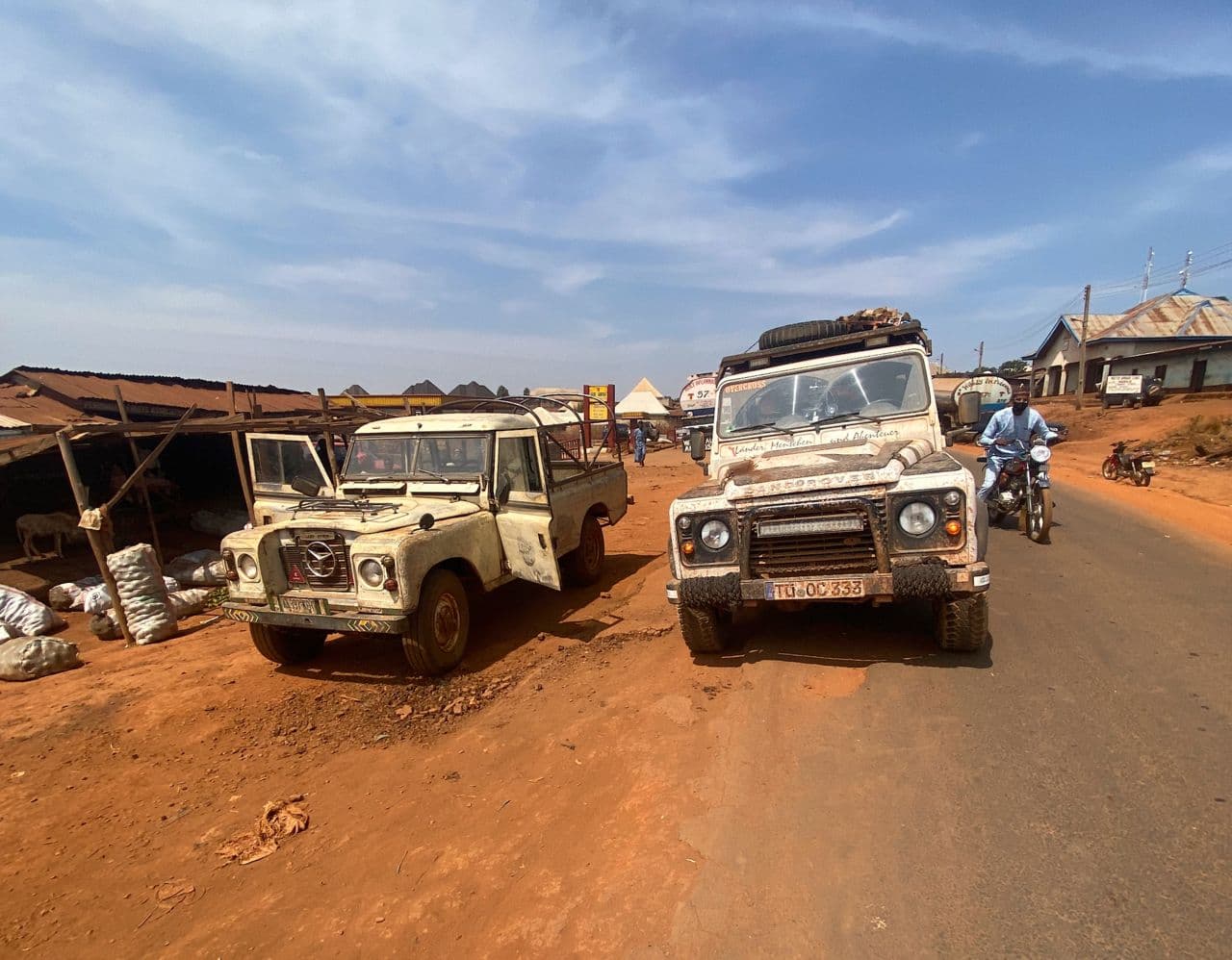 Dusty street scene with parked Land Rovers, a motorcyclist, market stalls, and red earth in a desert town.