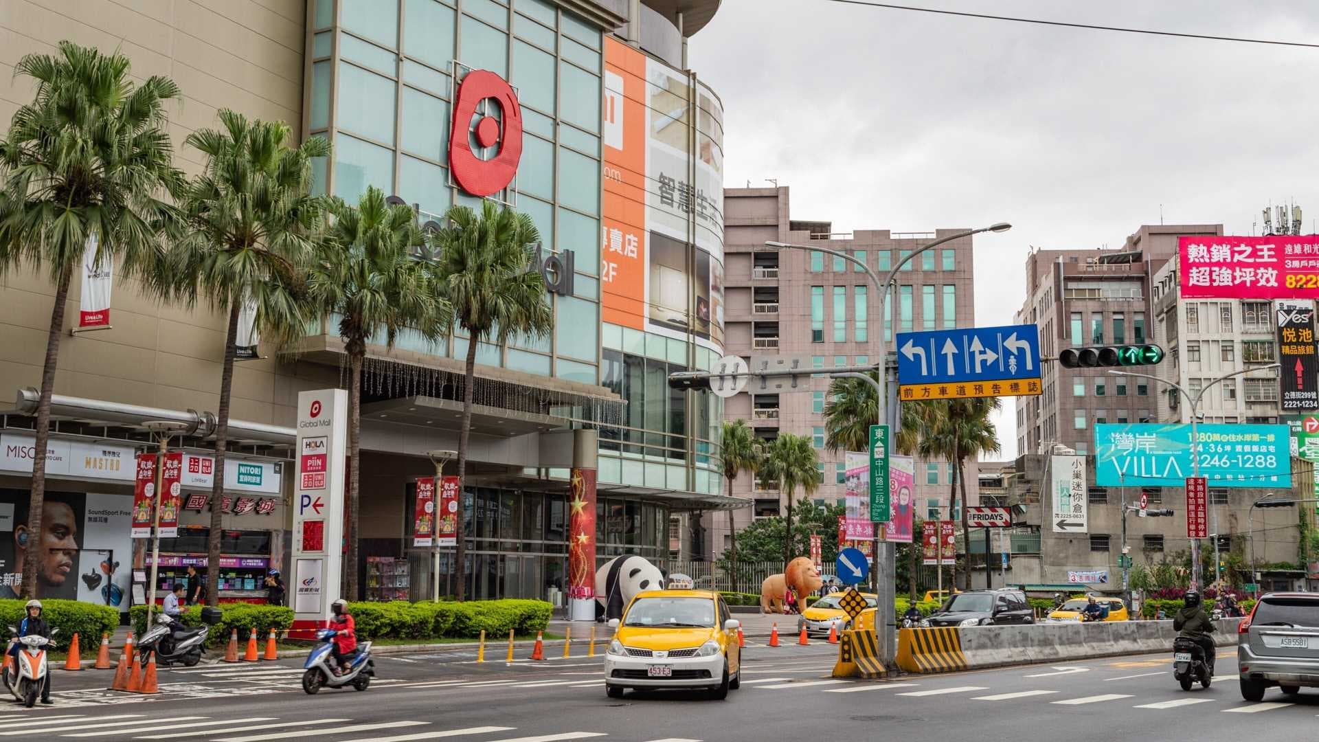 Busy urban streetscape featuring Global Mall in Banqiao, with palm trees, traffic, and commercial signage under an overcast sky.