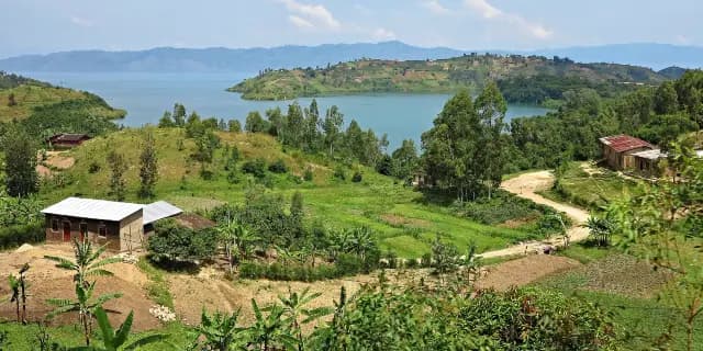 Wide landscape view of a river shoreline with rolling green hills, scattered houses, and distant mountains under a blue sky.