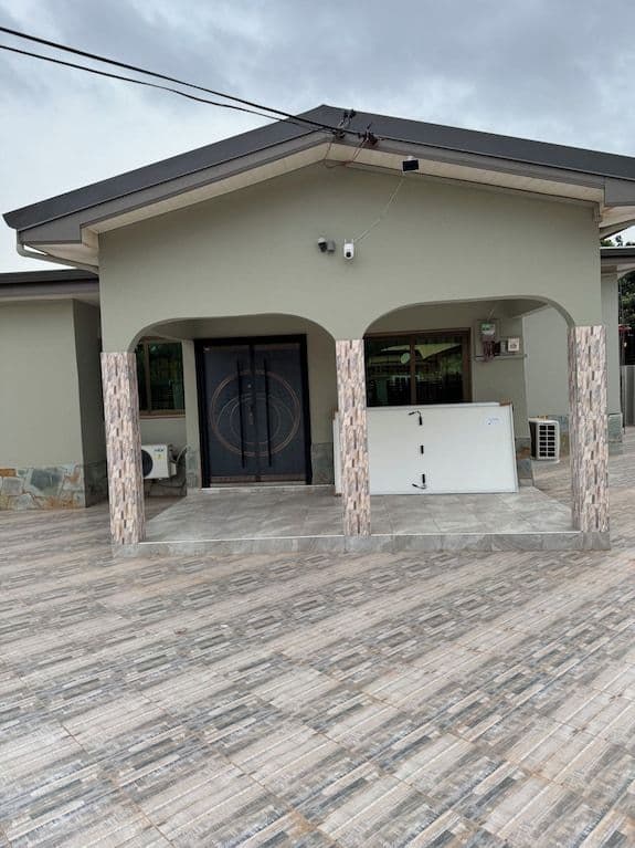 Exterior of a single-story residential house with a tiled driveway, arched porch, and security cameras under an overcast sky