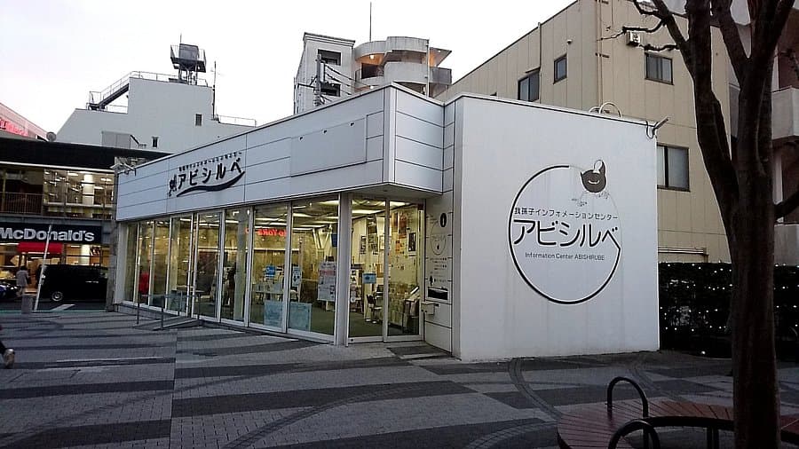 Exterior view of the white Abishure Information Center building with glass storefronts, situated on a paved plaza near a McDonald's.