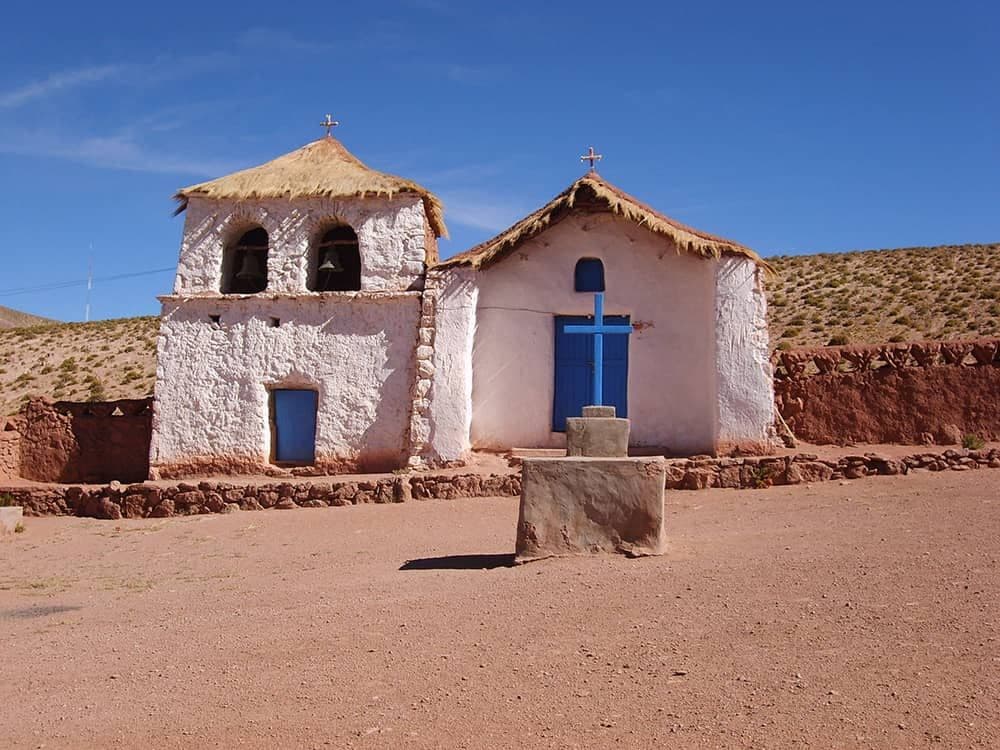 White adobe chapel with thatched roof and blue doors in a rural highland village setting.