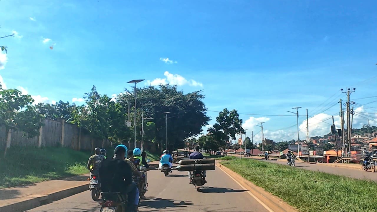 Motorcyclists transport heavy loads along a paved road in Bweyogerere, flanked by greenery and a hillside settlement under a bright blue sky.