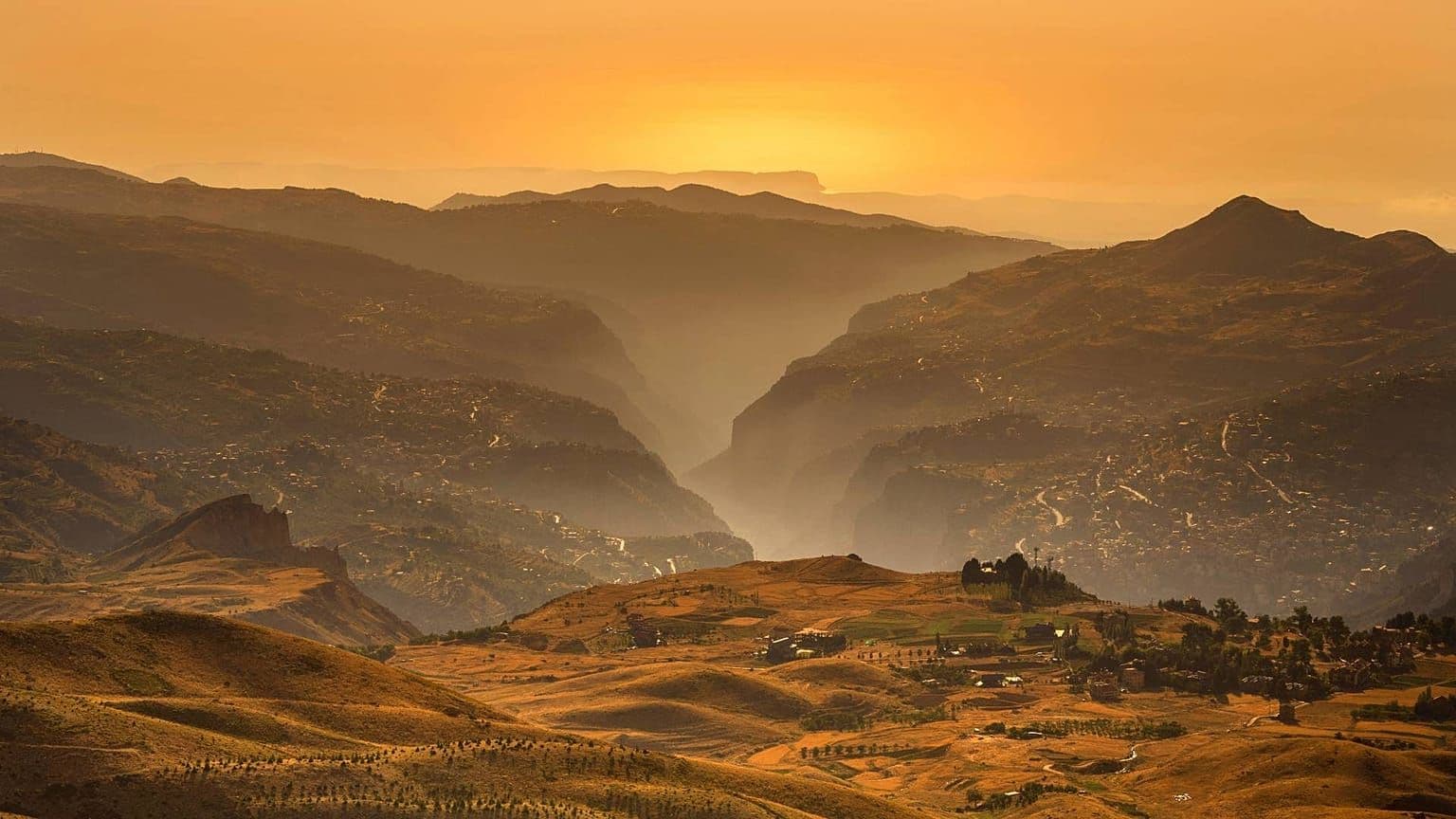 Golden-hour mountain landscape with layered ridges, hazy valleys, and warm autumn light over rolling hills.