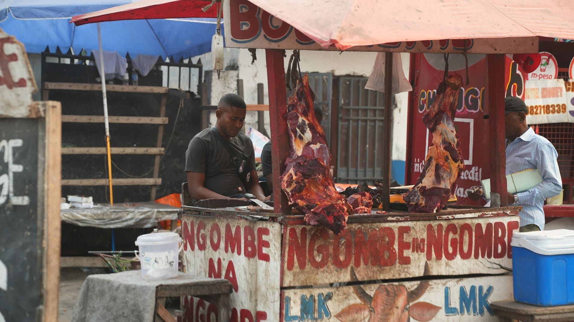 A street butcher stall in Bolobo displaying hanging raw meat carcasses, with a vendor working behind the counter under a makeshift shelter.