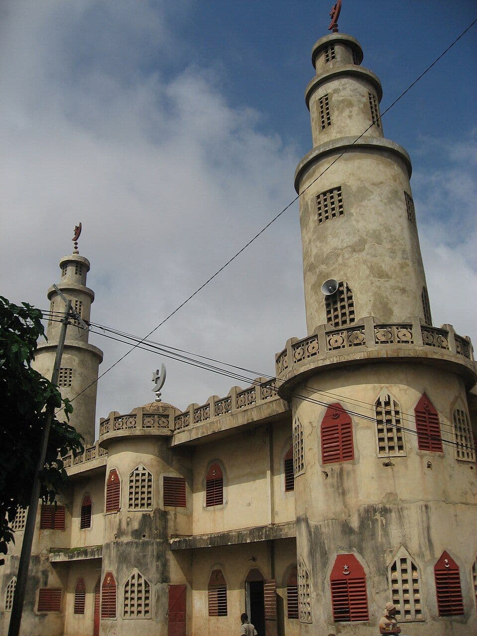Exterior of a mosque in the Borgou Department region, with two tall minarets, red-shuttered windows, and a weathered facade under a partly cloudy sky.