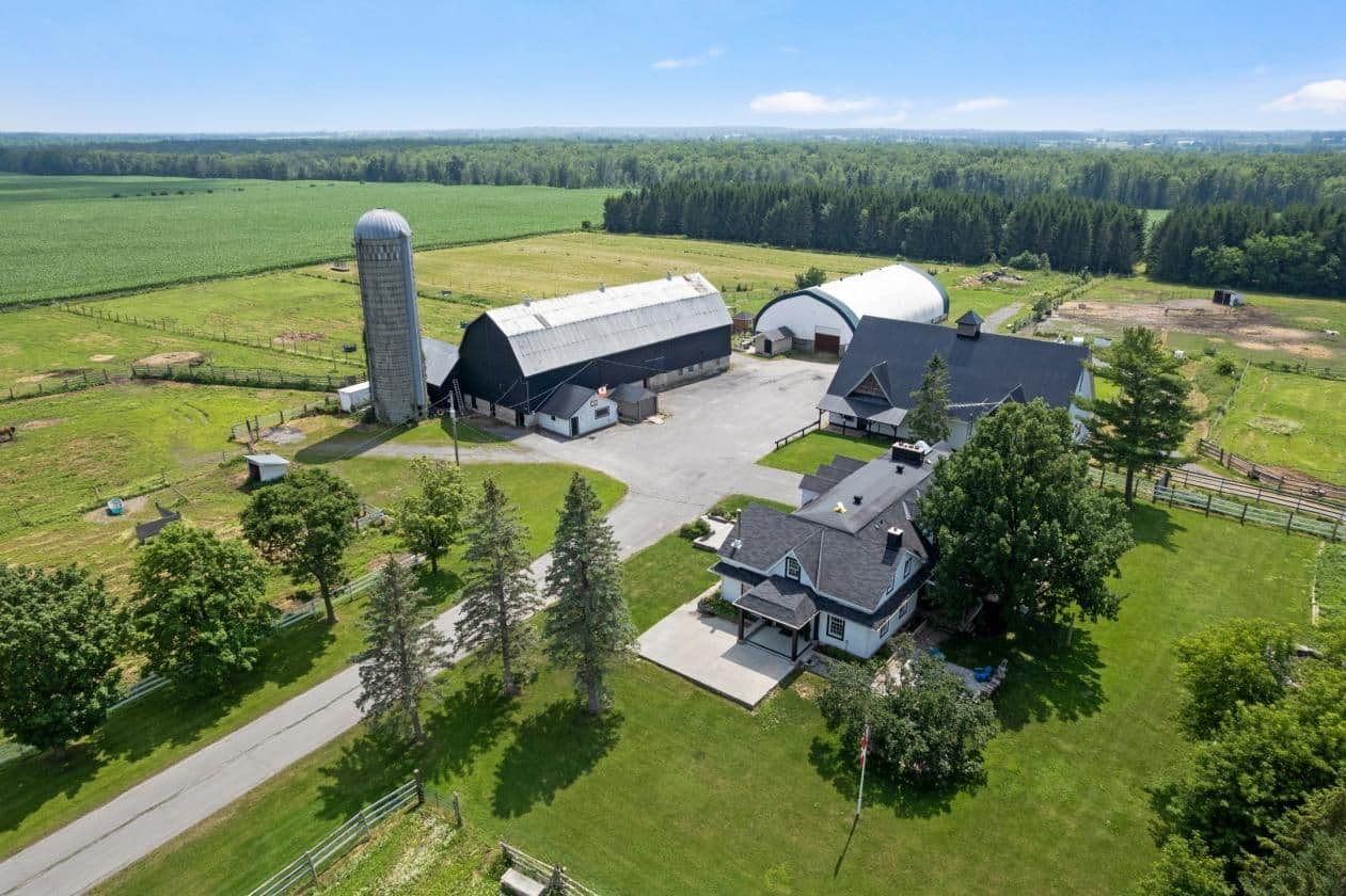 Aerial view of a rural farmstead with barns, silo, and farmhouse surrounded by green fields and forest.
