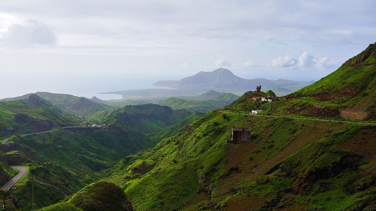 Winding road traverses lush green hills in the interior of Santiago Island, with distant mountains and coastline visible under a cloudy sky.