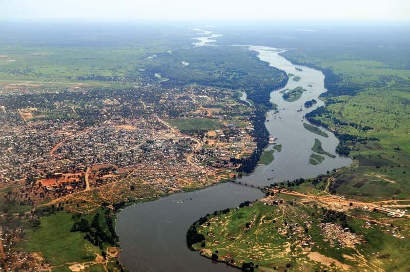 Aerial photograph of a wide river valley with a meandering waterway, green agricultural land, and a dense urban settlement on the left bank.