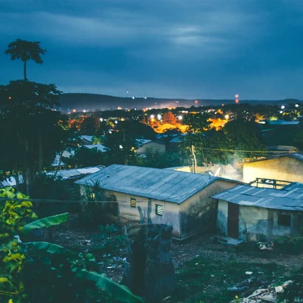 Twilight elevated view of Bassar town with corrugated-roof buildings, scattered lights, distant hills, and a tall palm tree silhouetted against a blue sky