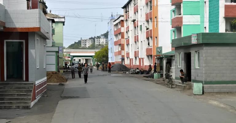 Wide street view of a North Korean residential settlement with colorful apartment blocks, small shops, and scattered pedestrians.