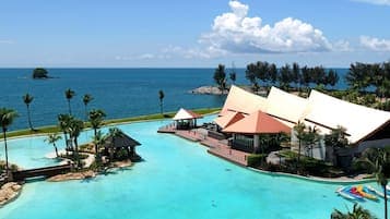 Aerial view of a tropical resort featuring a large swimming pool, thatched-roof buildings, and palm trees overlooking the blue ocean and coastline.