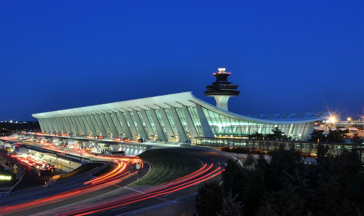 Nighttime long-exposure photograph of a modern airport terminal with control tower and light trails on surrounding roads.