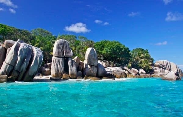 Coastal view of large granite boulders on a tropical shoreline with turquoise water and lush green vegetation under a blue sky.