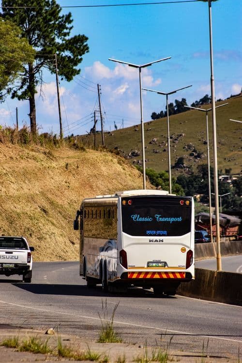 A white bus and pickup truck travel on a paved rural road through a hilly landscape near Sidvokodvo, Swaziland, under a blue sky.