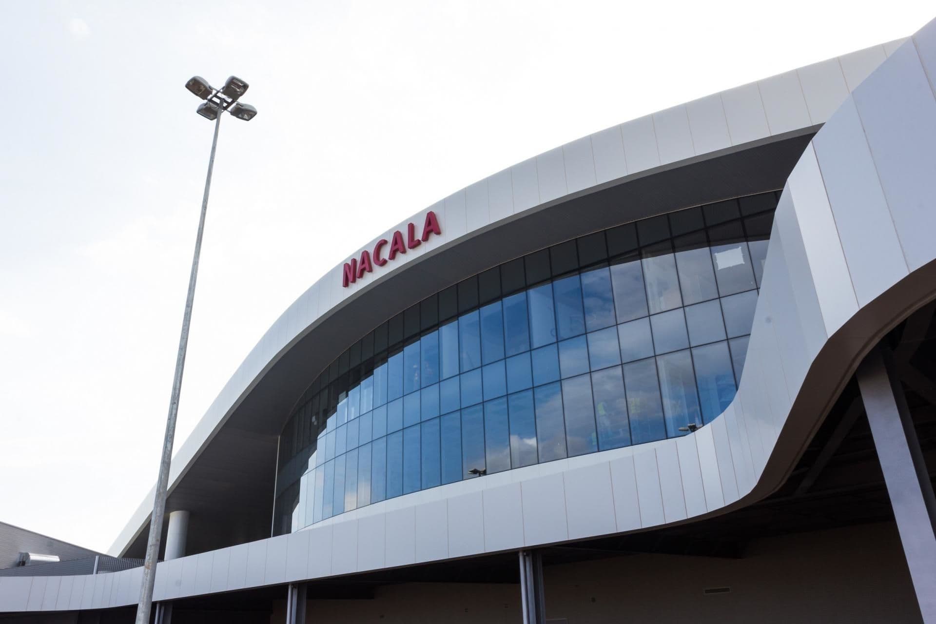 Low-angle shot of a modern white building facade featuring large blue glass windows and red 'NACALA' signage against a bright sky.