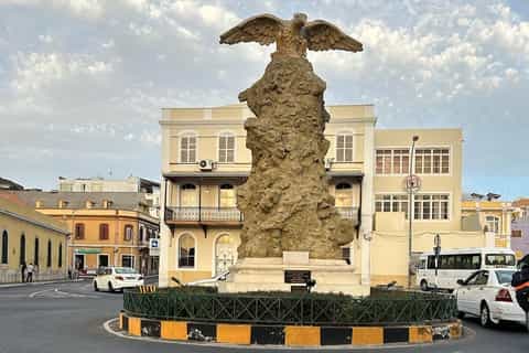 A large stone eagle monument stands on a traffic island in a town square, surrounded by low-rise buildings and passing vehicles under a cloudy sky.