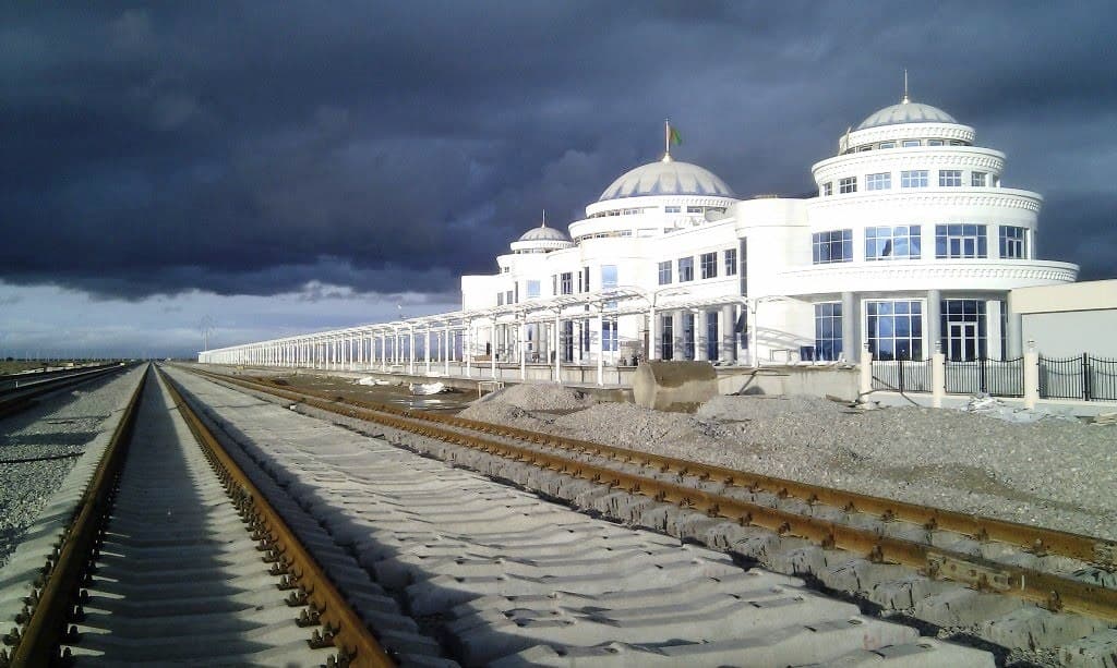 Exterior view of the white, domed Bereket Railway Station building under a dramatic cloudy sky, with railway tracks in the foreground.