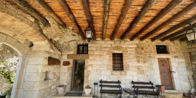 Traditional stone house facade with wooden ceiling beams, benches, and arched entryway in a village setting.