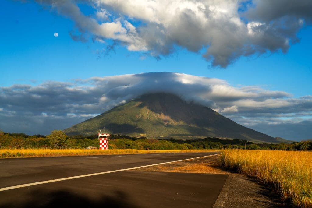 A paved road leads toward the Telica volcano under a blue sky with clouds and moon.