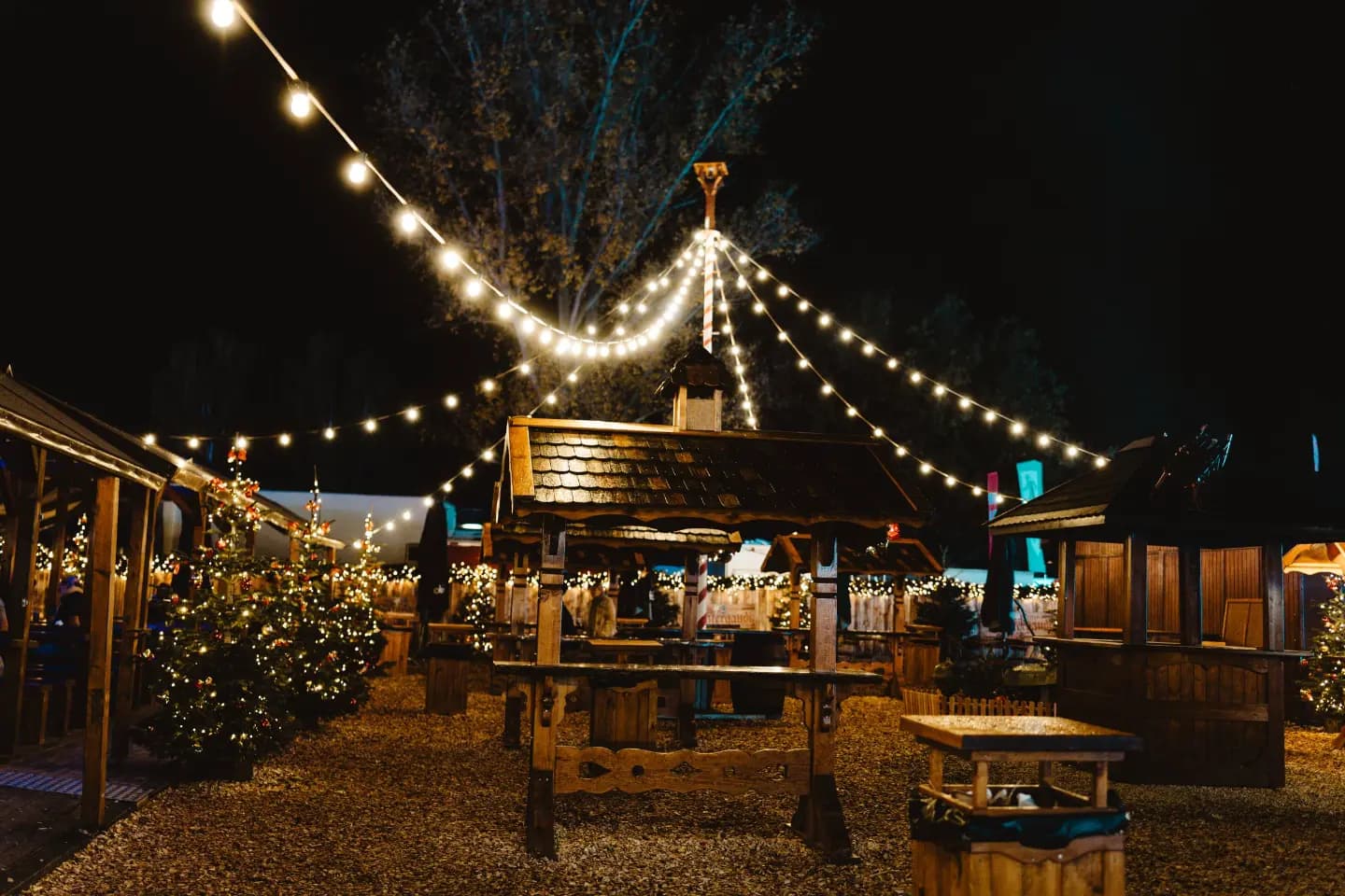 Nighttime open-air market at Tuzi with wooden stalls, string lights, and decorated trees.