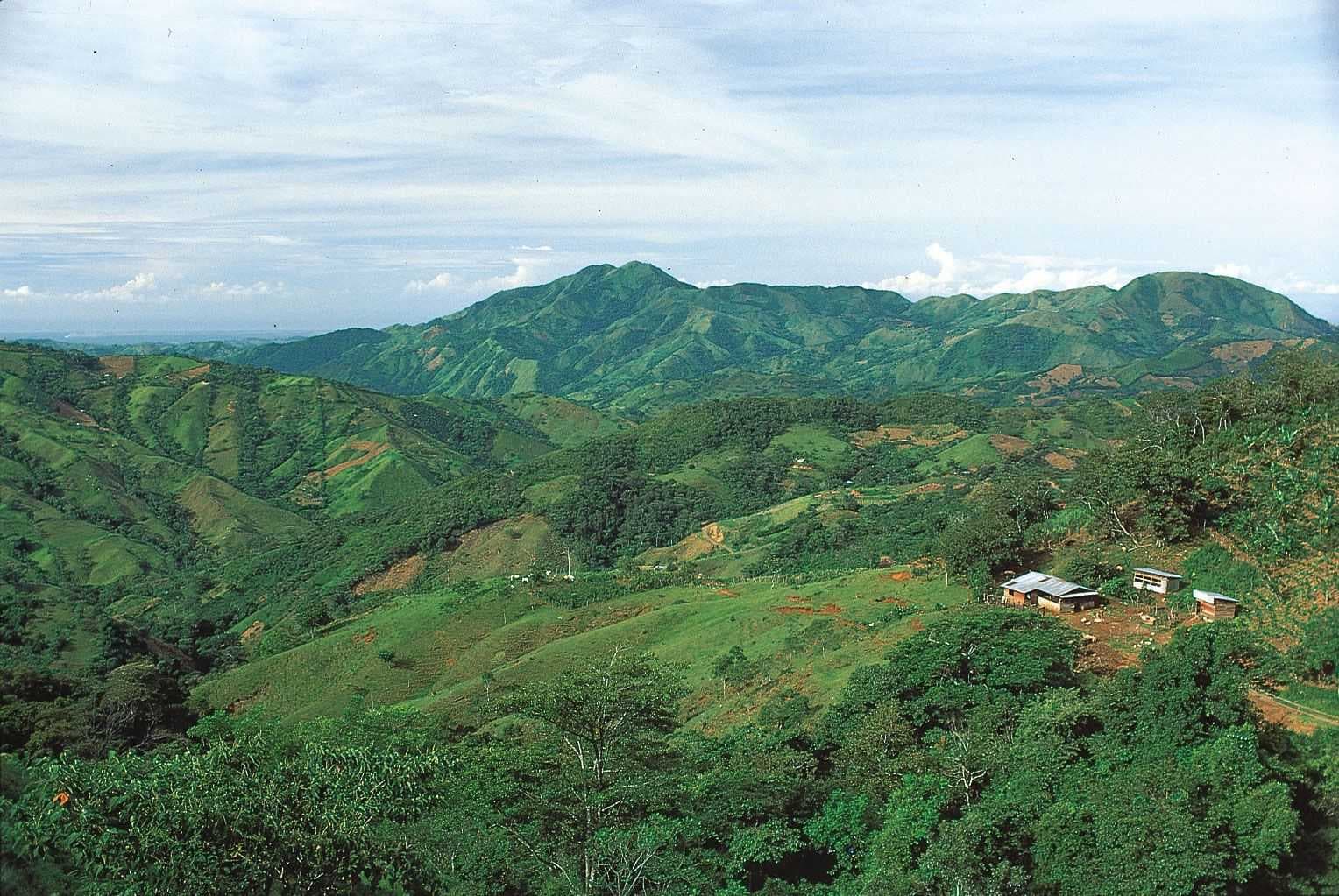 Wide landscape photograph of green rolling hills and mountains with scattered small buildings and vegetation under a partly cloudy sky.
