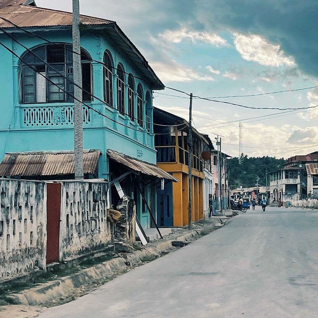 A quiet small-town street in Lindi, Tanzania, with colorful low-rise buildings, corrugated roofs, and overhead wires under a cloudy sky.