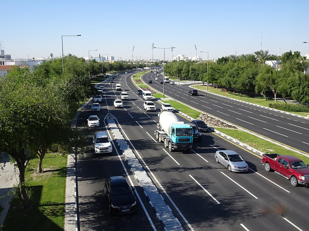 Aerial view of a multi-lane highway with moderate traffic, including a cement mixer and red pickup, flanked by green trees under a clear blue sky.