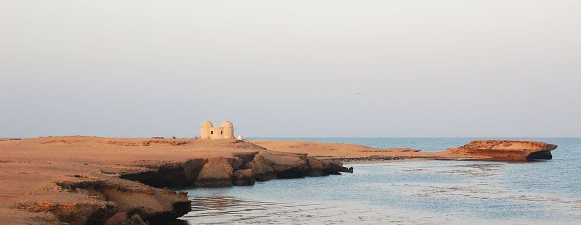 Wide coastal landscape of arid sand dunes, rocky shoreline, and small domed structures near the sea on Kamaran Island, Yemen.