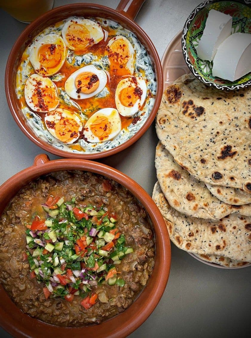 Overhead shot of Ful Medames with eggs, salad, and flatbreads in terracotta bowls on a metal table.
