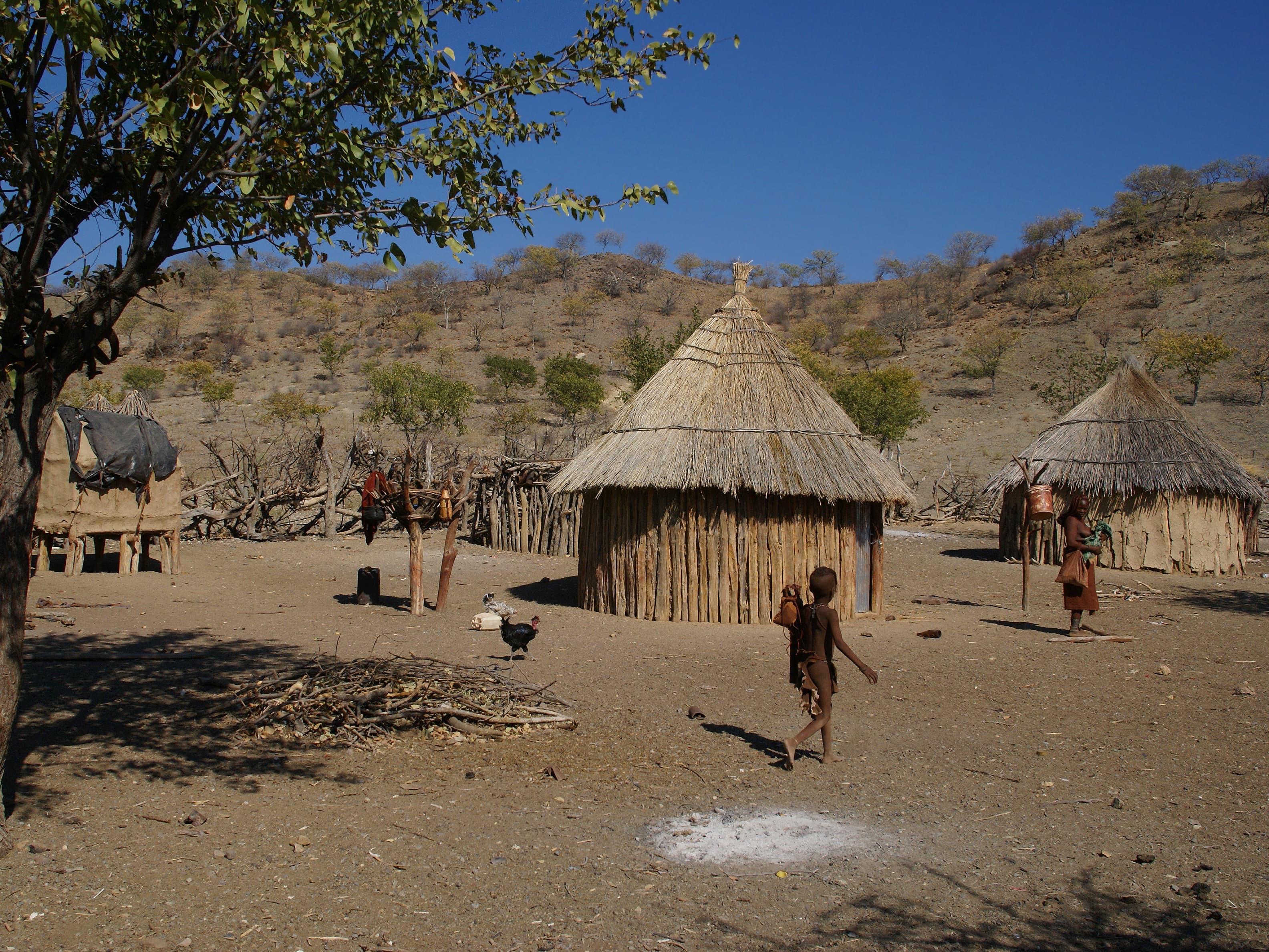 A rural village scene featuring traditional thatched huts, a child walking across dry earth, and arid hills under a clear blue sky.