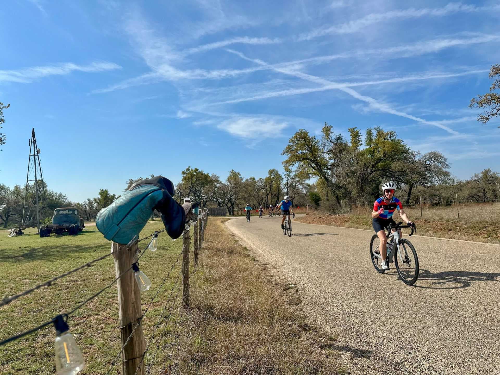 Cyclists ride a paved rural road under a blue sky, with a fence, grass, and trees in the foreground