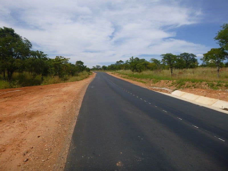 A paved two-lane road stretches through a rural landscape with red dirt shoulders, scattered trees, and a partly cloudy sky.