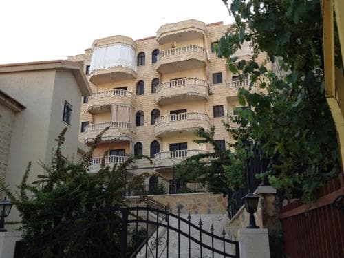 Exterior view of a multi-story beige residential building with curved balconies, framed by green foliage and a black metal gate in the foreground.