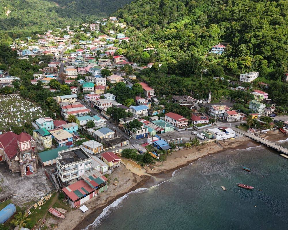 Aerial view of a colorful coastal village in Dominica with a church, beach, and boats along a lush, hilly shoreline.