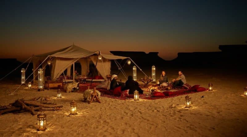 A group of people dining on red carpets in a desert setting at dusk, illuminated by lanterns near a large luxury tent.