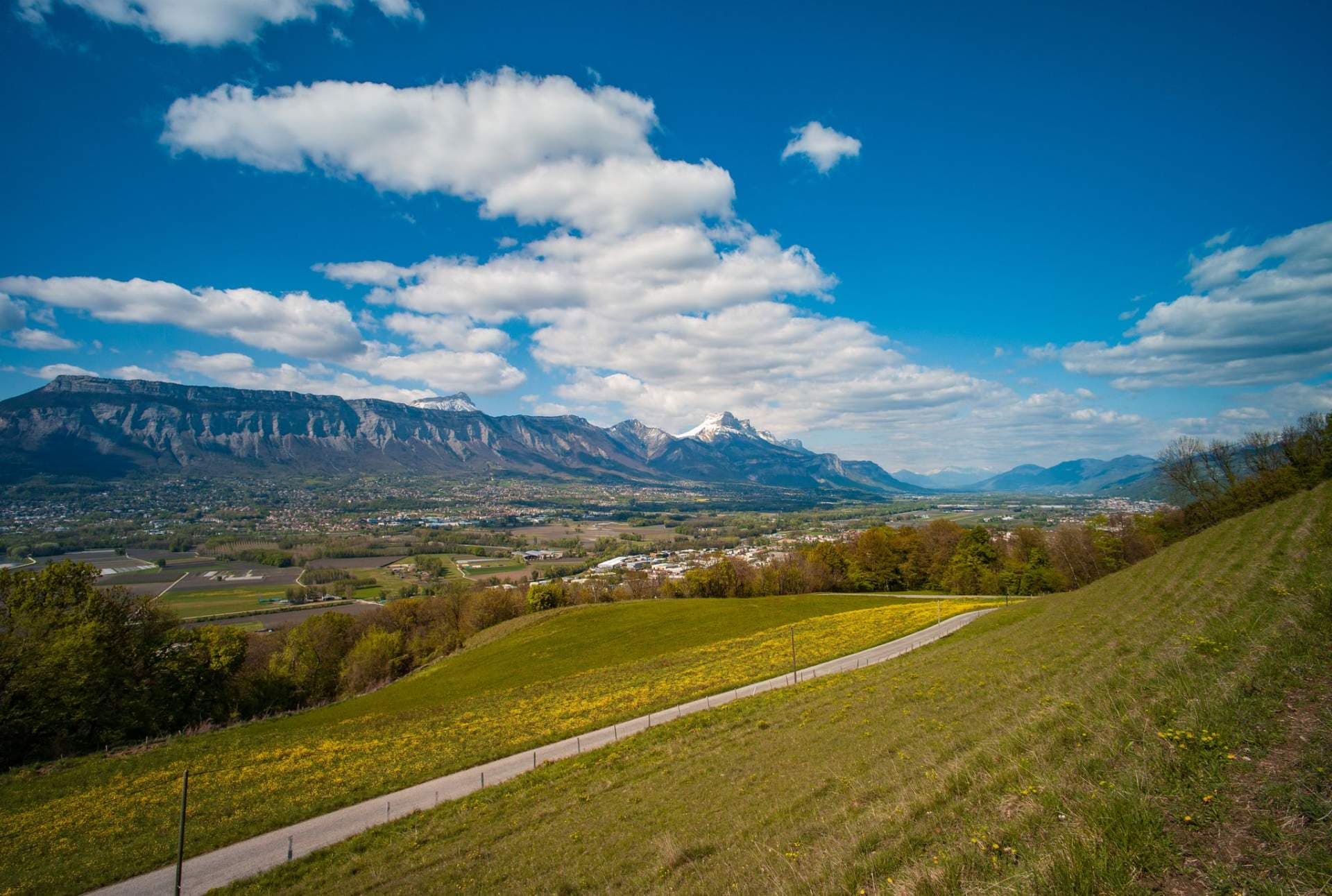 Grassy hillside path with wildflowers leading toward a distant town and limestone cliffs near Saint-Cassien, France