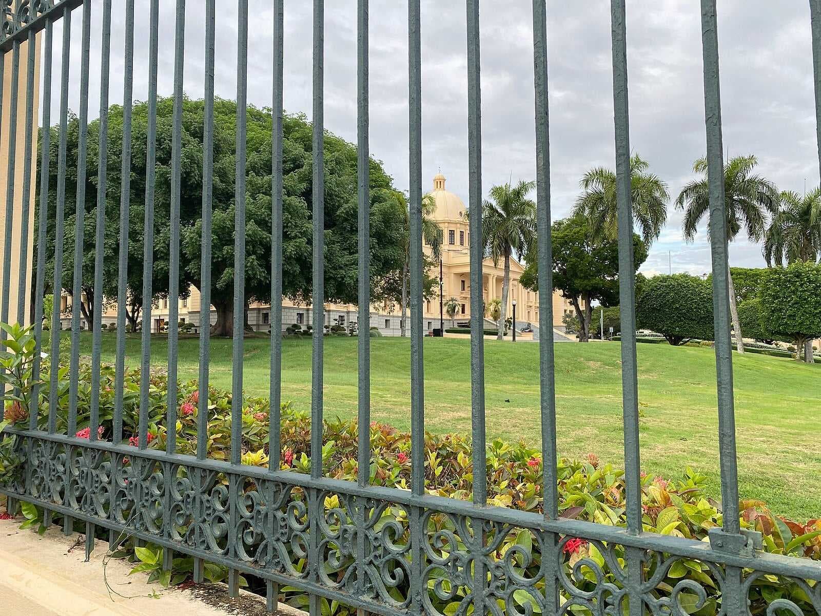 View through a green iron fence of a manicured lawn, tropical trees, and a domed colonial-style building under an overcast sky near Bisonó.