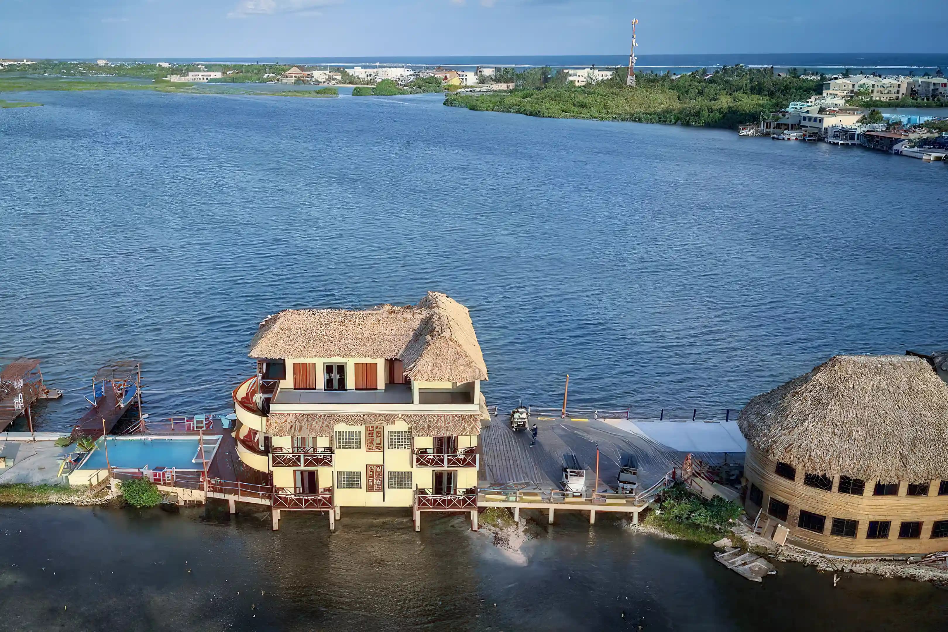 Aerial view of a waterfront resort with thatched-roof buildings, a swimming pool, and a dock extending into a wide blue river channel.