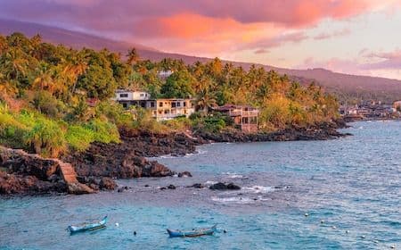 Tropical coastline with turquoise water, small boats, rocky shore, lush vegetation, and buildings under a sunset sky.