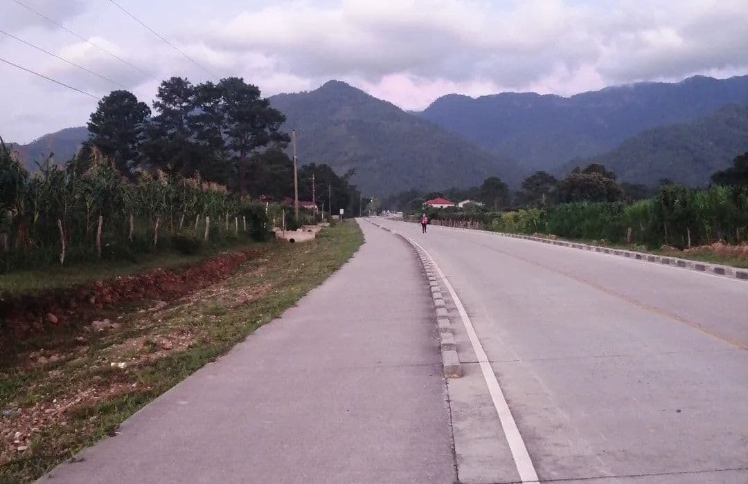A paved rural road stretches toward distant green mountains under a cloudy sky, flanked by vegetation and small structures.