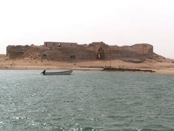 Coastal view of a rocky island with ancient stone ruins and a small boat on the water near the Kamaran shoreline.