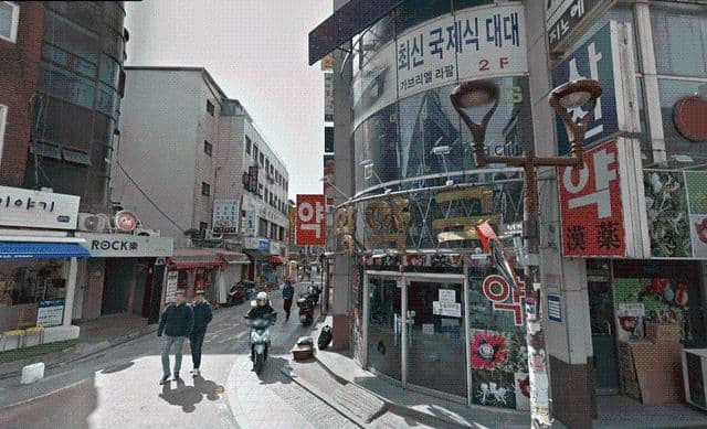 Narrow shopping street with independent shops, Korean signage, pedestrians, and a scooter rider in an urban commercial district.