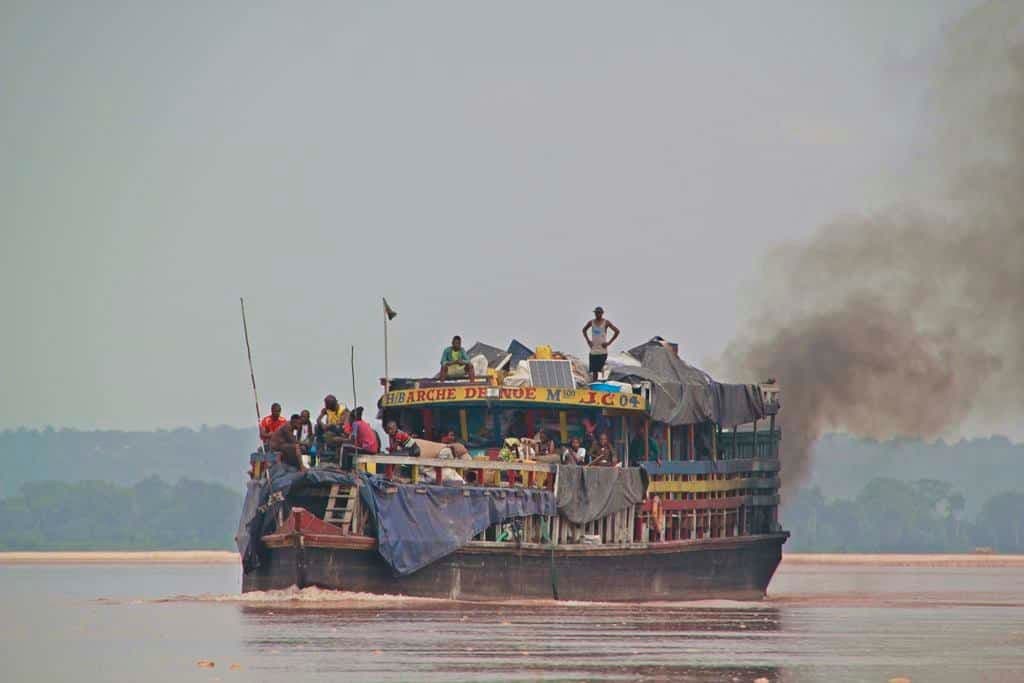A crowded river boat emitting black smoke travels across a wide river, likely the Congo, with passengers visible on deck.