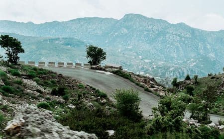 A winding paved road curves through a rugged, mountainous landscape with sparse vegetation and distant peaks under an overcast sky.