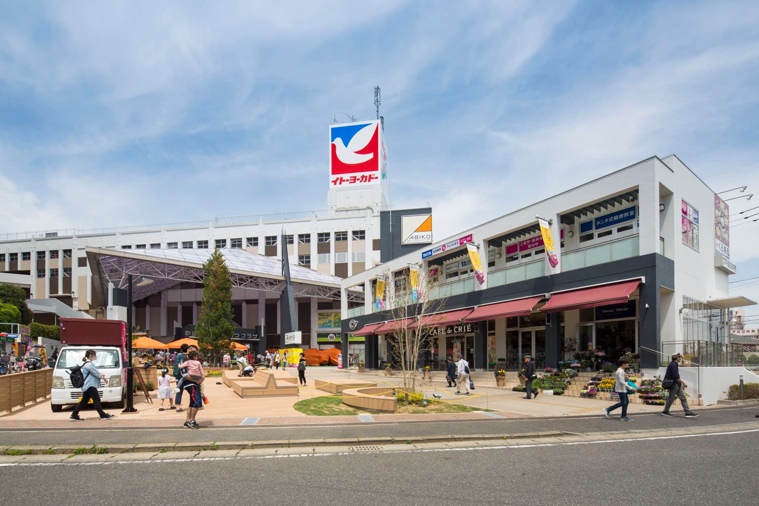 Exterior view of the Abiko Station building featuring the Ito-Yokado sign, a plaza with pedestrians, and adjacent commercial storefronts under a blue sky.